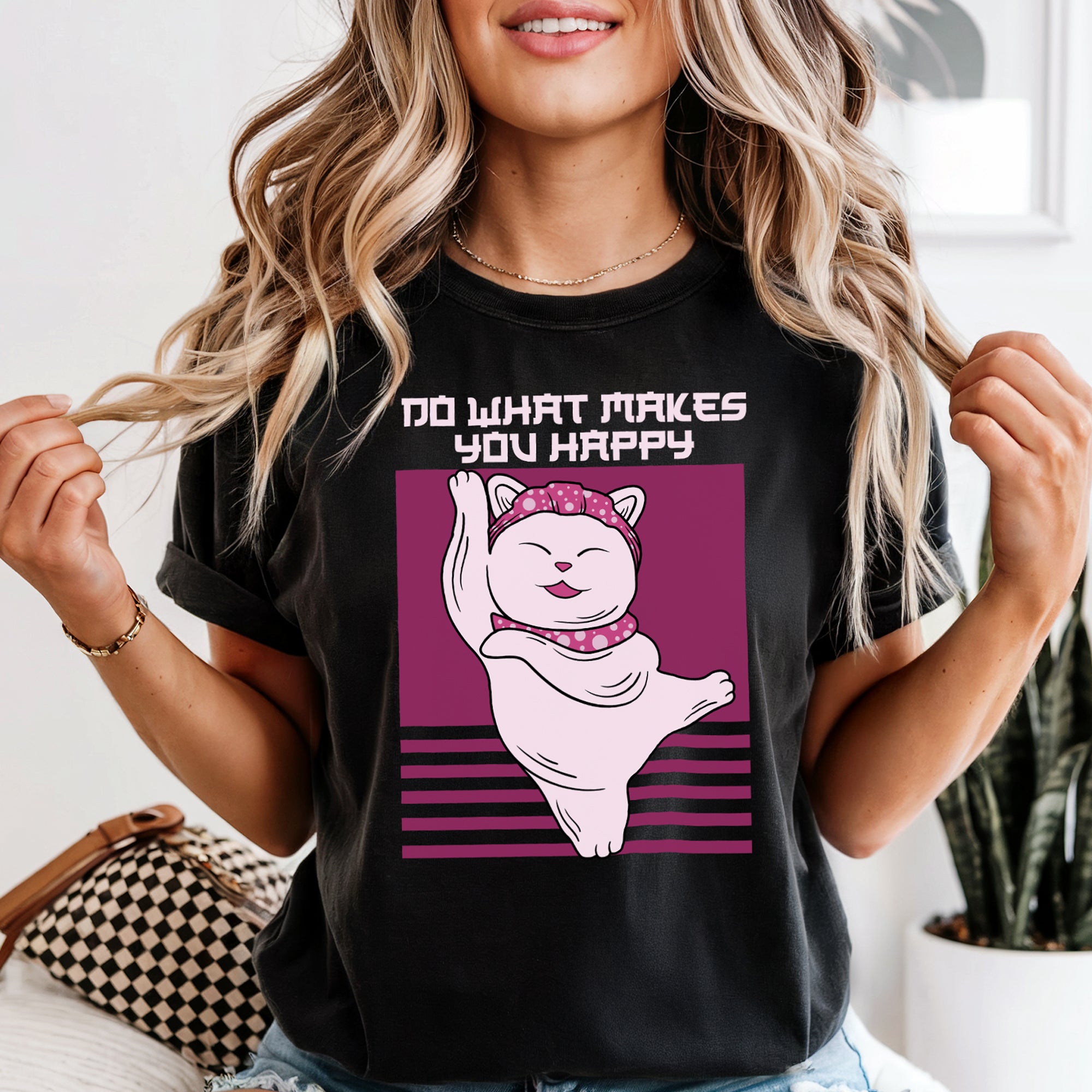 A female model wearing the black Vintage Ballet Cat Do What Makes You Happy Women's Graphic Tee, featuring a cat in a pink headband and scarf performing a graceful dance move. Model is sitting on a couch.