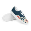 A pair of women's Orca Snork canvas sneakers, featuring a navy blue and floral pattern, are shown from a right-side product shot. The top shoe is resting on its sole, propped up to reveal the bottom tread of the other shoe. The shoes have a dark navy blue upper and a cream-colored lower with a pink and blue floral design. The white laces and soles provide a sharp contrast.