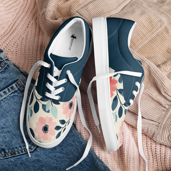 A flat lay product shot of a pair of women's Orca Snork canvas sneakers. The shoes are resting on a background of light pink ribbed sweater fabric and a section of blue denim. The sneakers have a dark navy blue upper and a cream-colored lower with a pink and blue floral pattern. The white laces are loosely tied and trail across the background.